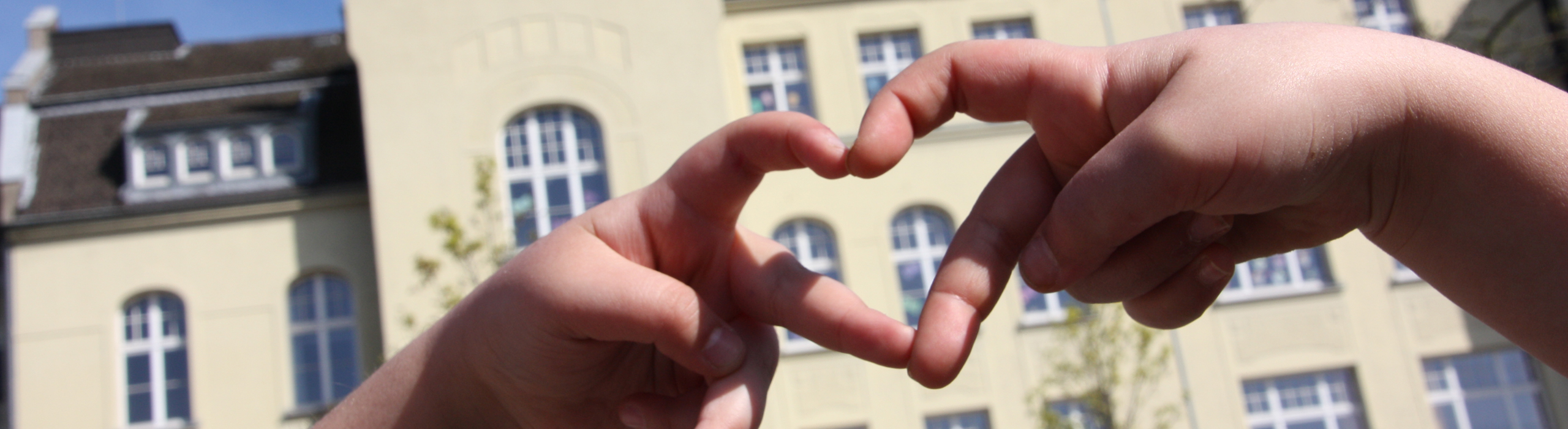Zu einem Herz geformte Hände vor dem Gebäude der Gemeinschaftsschule Stoffeler Straße